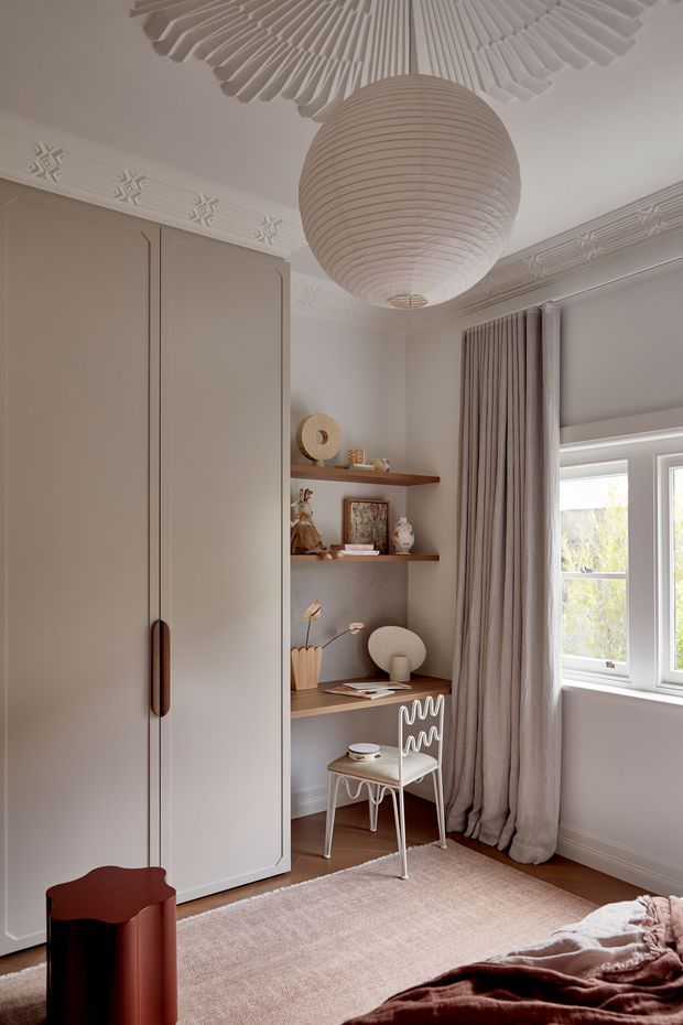 A refined, compact bedroom corner featuring a built-in wardrobe, integrated desk and open shelving, a small sculptural chair, and a partially visible bed. The room has warm taupe walls, ornate white ceiling cornices and decorative ceiling detailing, timber flooring, full-height neutral curtains, and a large ribbed paper pendant light. A soft neutral rug and carefully styled shelving create a calm, sophisticated atmosphere with subtle Art Deco-inspired detailing.