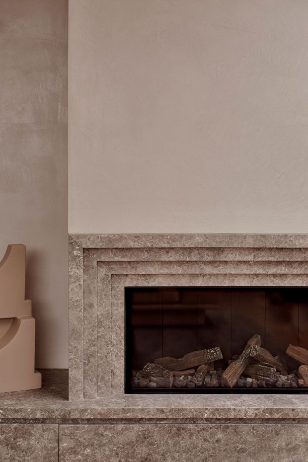 Close-up interior view of a contemporary fireplace with a broad, layered natural-stone surround and hearth. The walls are finished in warm taupe textured plaster, while a dark inset firebox contains decorative logs and stones. A sculptural neutral-toned object is partially visible at the left edge. The composition is warm, earthy, refined, and focused on the fireplace feature.