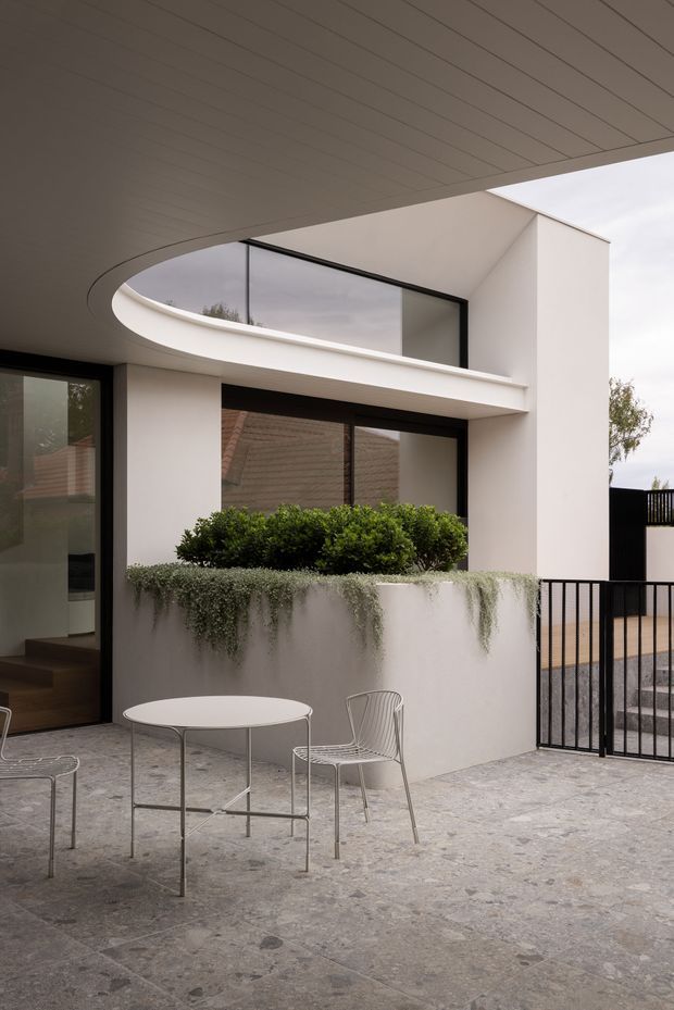 A refined modern courtyard or terrace attached to a white rendered house. The space features pale stone paving, a curved overhead soffit, black-framed glazing, a raised planter with clipped shrubs and trailing greenery, and a minimalist round outdoor table with wire chairs. A black metal gate and steps define the edge of the courtyard.
