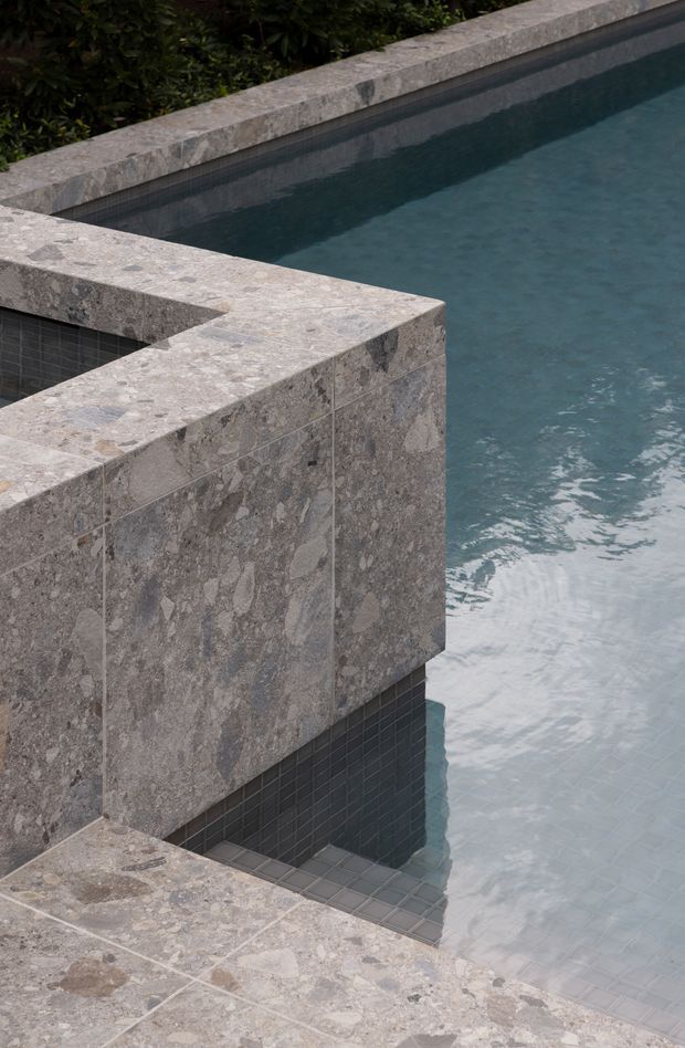 Close-up architectural view of a contemporary swimming pool with dark blue-grey tiled walls and water, framed by substantial grey natural-stone coping and tiled surfaces. A raised or projecting stone-clad pool structure creates a geometric, sculptural composition, with dense greenery visible beyond the pool edge.