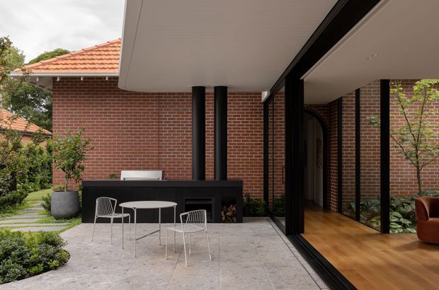 A contemporary courtyard and outdoor entertaining area beside a brick residential extension. The space features a paved terrace with a black outdoor kitchen or barbecue enclosure, small round dining table and wire chairs, raised planting, and a covered ceiling. Full-height black-framed glazing connects the terrace to a warm timber-floored interior, while the original red-brick house and terracotta tiled roof remain visible beyond.