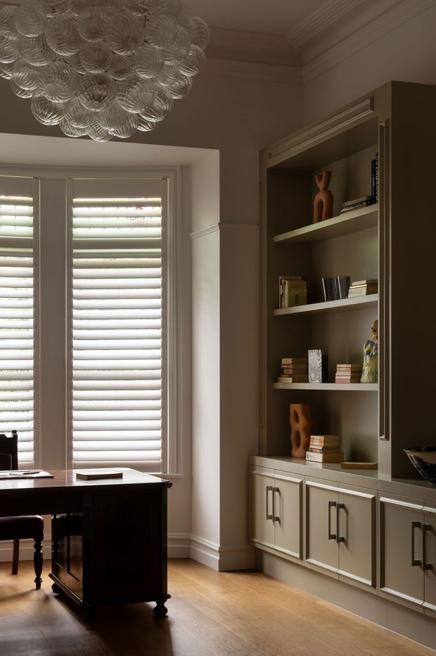 Elegant traditional home office with a dark wood desk positioned beside tall plantation-shuttered windows. The room features warm timber flooring, taupe painted walls with substantial moulded cornices and trim, and a large built-in bookcase with lower cabinet storage in a muted grey-green finish. A sculptural glass pendant light adds a decorative focal point overhead.