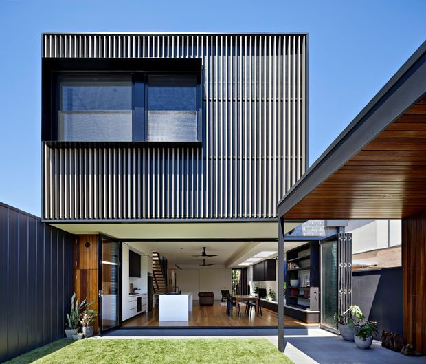 Exterior view of a contemporary multi-level residence centered on a private courtyard. The upper storey is wrapped in dense vertical slatted screening with a projecting dark-framed window, while the ground level opens broadly to the courtyard through large glazed doors. A timber-lined covered outdoor area and dark metal cladding frame the right side, with the interior kitchen, dining and living spaces visible beyond.