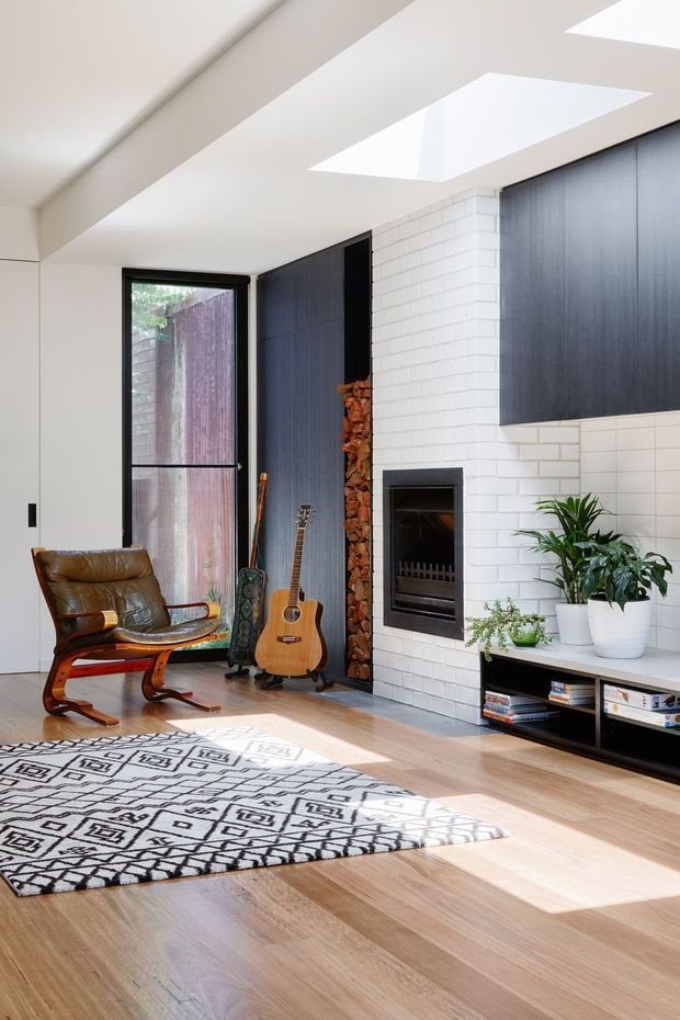 Bright contemporary living room featuring a white-painted brick fireplace with integrated black firebox, dark built-in cabinetry, an open firewood niche, skylights, light timber flooring, a leather lounge chair, guitar, indoor plants, and a black low media console. A patterned monochrome rug anchors the seating area.