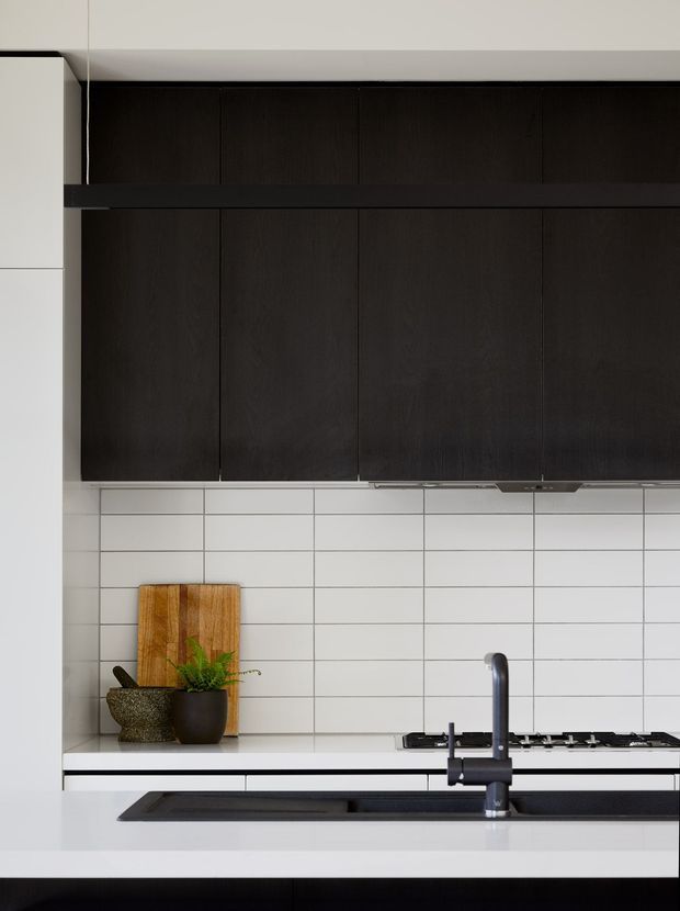 A compact, highly designed kitchen featuring dark wood or near-black upper cabinetry, white tiled splashback with dark grout, and a white benchtop. A black undermount sink with a tall dark mixer tap sits in the foreground, while a black gas cooktop is positioned along the rear counter. A timber chopping board and potted plant add warmth against the restrained monochrome palette.