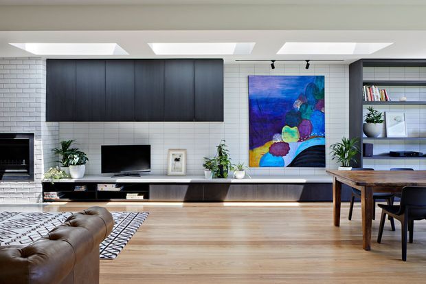 A bright, contemporary open-plan living and dining area with a long built-in entertainment wall, dark timber cabinetry, white brick and tiled wall finishes, and a large colourful artwork. The room includes a fireplace, television, low media joinery, timber flooring, a leather sofa, patterned rug, timber dining table, open shelving, indoor plants, and multiple skylights.