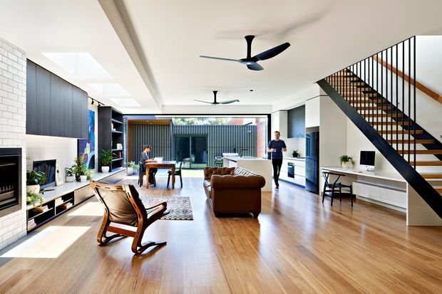 Bright open-plan contemporary living space with polished timber flooring, white painted brick and plaster walls, extensive glazing, and a dramatic black open-riser staircase with slim vertical balustrade. The room includes a fireplace and built-in media joinery on the left, lounge seating in the centre, a dining area toward the rear, and kitchen cabinetry on the right. Skylights, ceiling fans, and a view toward a screened outdoor courtyard add to the airy, indoor–outdoor character.