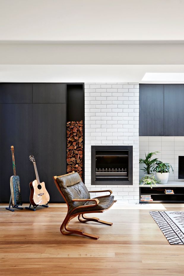 A bright contemporary living room featuring a full-height dark timber media and storage wall, open firewood niche, white brick fireplace surround, built-in black fireplace, and low entertainment cabinetry. A leather sling lounge chair, guitars, potted plants, books, and a patterned rug add warmth and character. Natural timber flooring and a skylight contribute to the airy atmosphere.