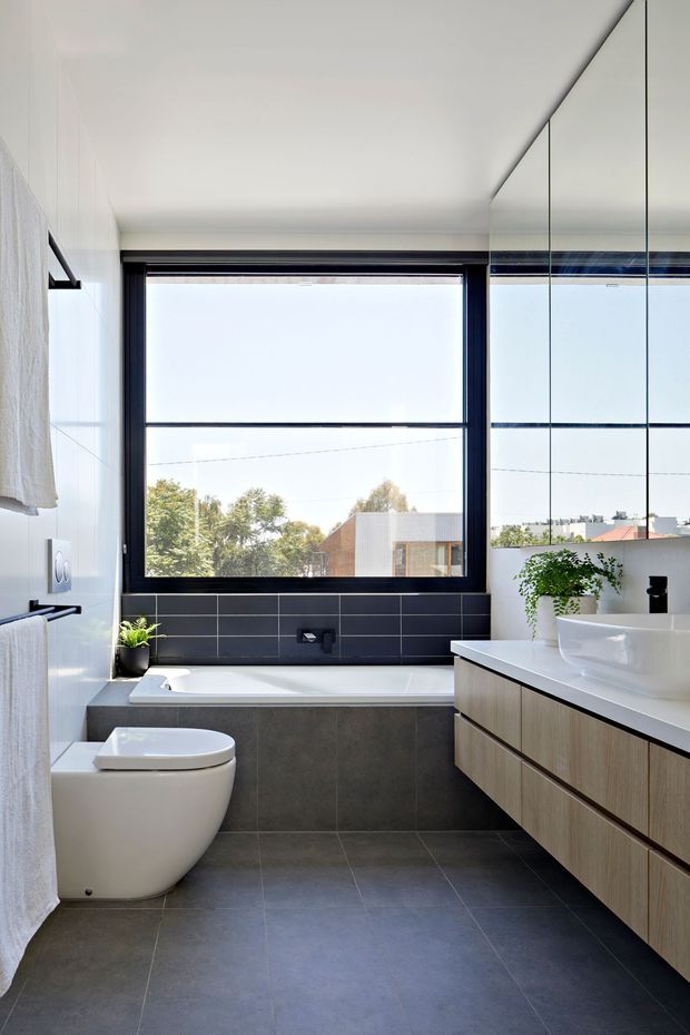 A bright, contemporary bathroom with a freestanding-style bathtub beneath a large black-framed window, a wall-hung-style white toilet, and a long timber-look floating vanity with an integrated white basin. Dark grey floor tiles and matching horizontal wall tiles contrast with white walls and mirrored upper cabinetry. A small indoor plant adds greenery, while neighbouring residential buildings and trees are visible through the window.