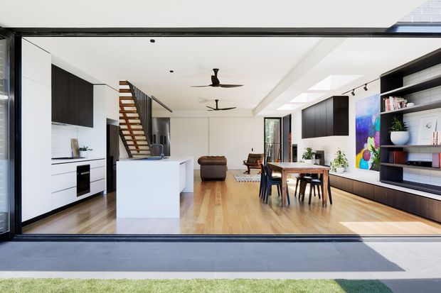 Bright, open-plan contemporary interior viewed through fully opened glazed doors, combining a white kitchen with island, dining area, living space, floating timber stair, built-in dark shelving, skylights and ceiling fans. The room has light timber flooring, crisp white surfaces and strong indoor-outdoor visual connection.