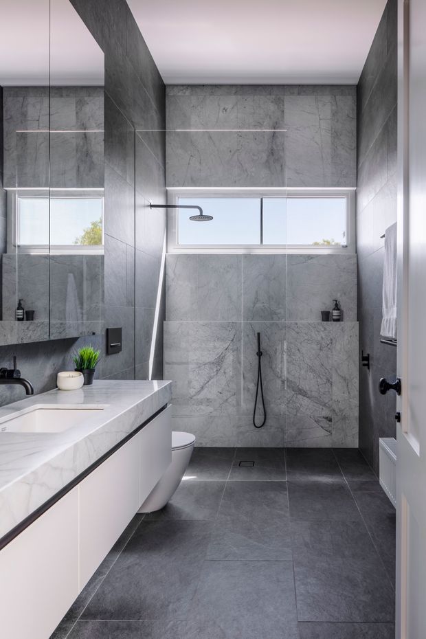 A contemporary bathroom with full-height grey marble-look wall tiling and matching dark grey floor tiles. The room features a frameless glass shower enclosure, wall-mounted black shower fittings, a floating white vanity with a pale stone benchtop, integrated basin, wall-hung toilet, horizontal clerestory window, and large mirrored panel. The palette is monochromatic grey, white, and black with clean minimalist detailing.