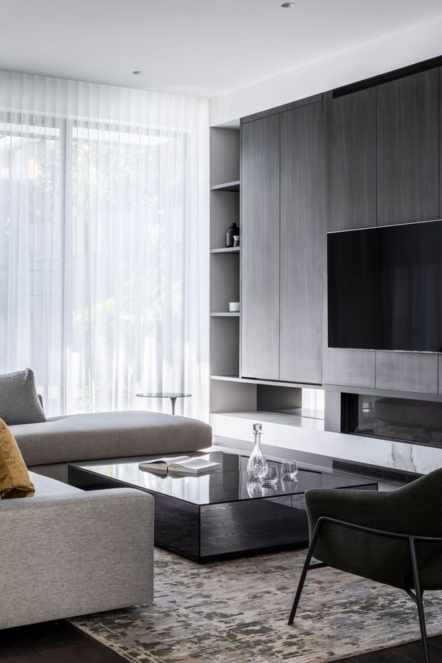 A refined contemporary living room with a large grey sectional sofa, dark upholstered lounge chair, black reflective coffee table, and textured area rug. A full-height dark timber-toned built-in media wall incorporates open shelving, a wall-mounted television, ventilation, and a linear fireplace. Sheer white curtains cover large windows, while recessed ceiling lights and pale walls create a bright, polished interior.