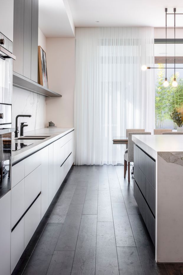 Bright contemporary kitchen with a long run of white flat-panel cabinetry, integrated appliances, dark flooring, and a white marble-look splashback. A dark island with a pale stone benchtop occupies the right side, opening toward a dining area with upholstered chairs. Full-height sheer curtains and large glazing bring in abundant natural light, complemented by slim black-framed pendant lighting.