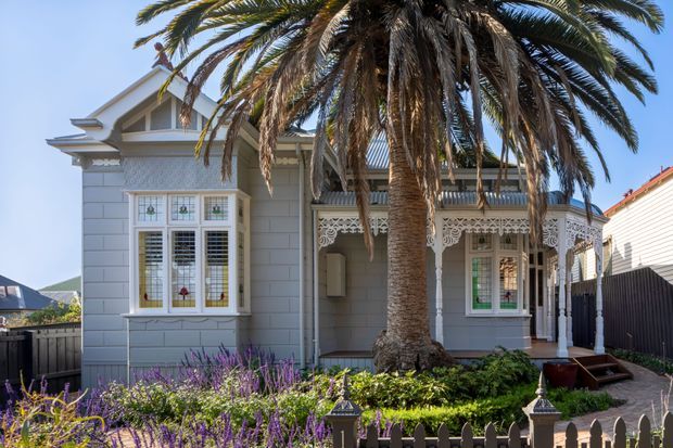 Front exterior view of a historic single-storey residence with pale grey weatherboard cladding, ornate timber verandah detailing, decorative window trim, and a steep gabled roof. A large palm tree and established garden occupy the foreground, with a low picket fence and paved path leading to the entrance.
