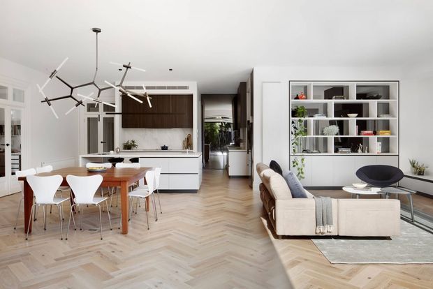 Bright open-plan contemporary interior combining a dining area, kitchen and living room. The space features herringbone timber flooring, white walls and cabinetry, a dark timber kitchen feature, a built-in open display shelving unit, modern furniture, and a sculptural branching pendant light.