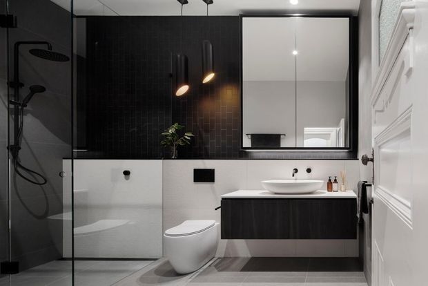Contemporary bathroom with a black and white scheme, featuring a glass-enclosed shower, freestanding vessel basin, wall-mounted dark timber vanity, wall-hung toilet, large mirror, and contrasting white decorative door and wall mouldings. Black tiled feature walls and pendant lighting add a refined, dramatic character.