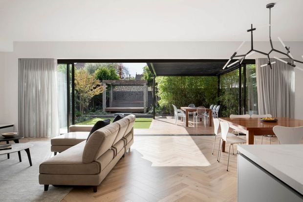 Bright open-plan contemporary living, dining and kitchen space with floor-to-ceiling glazing opening onto a landscaped outdoor area. The room features a pale herringbone timber floor, cream sectional sofa, white curtains, a timber dining table with white chairs, white kitchen cabinetry and a sculptural black-and-white pendant light. A covered outdoor dining area and garden are visible beyond the glazing.