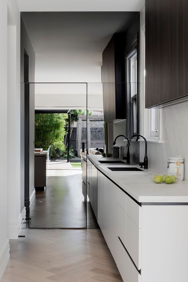 A narrow, contemporary galley kitchen viewed lengthwise toward a glazed opening and green courtyard. The kitchen features white flat-panel base cabinetry, dark wood-grain full-height cabinetry, a pale stone benchtop and splashback, black tapware and sink, integrated appliances, and light timber herringbone flooring. A large mirror at the end of the kitchen reflects the adjoining space and garden beyond.
