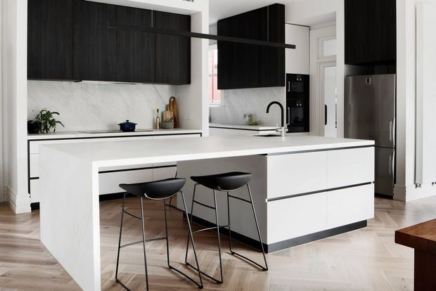Contemporary kitchen with a large white waterfall island and integrated seating, dark wood cabinetry, white marble-look splashback, stainless-steel refrigerator, and light timber flooring. Black pendant lighting and bar stools provide contrast in the bright, refined space.