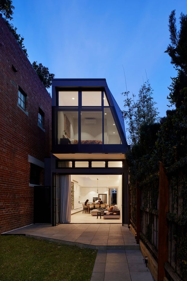 Twilight view from a small rear garden toward a narrow, contemporary multi-storey residential extension. The lower level opens through large glazed doors onto a paved terrace and reveals a bright living and dining area, while the upper level has expansive dark-framed glazing and a visible bedroom or sitting space. A red-brick wall borders the left side, with timber fencing and dense planting on the right. The extension features strong rectilinear lines, dark metal-framed windows, concealed lighting, and a projecting flat roof.