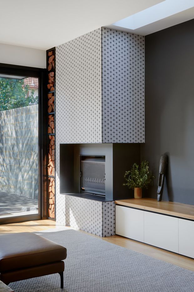 A refined contemporary living room featuring a built-in media and storage wall with striking black-and-white geometric patterned panels, a recessed fireplace or heater, and low white cabinetry topped with a timber surface. A large glazed door brings in sunlight beside a vertical stacked timber-log feature. The room includes a neutral grey carpet, dark leather seating, a potted plant, sculptural decor, and a linear skylight.