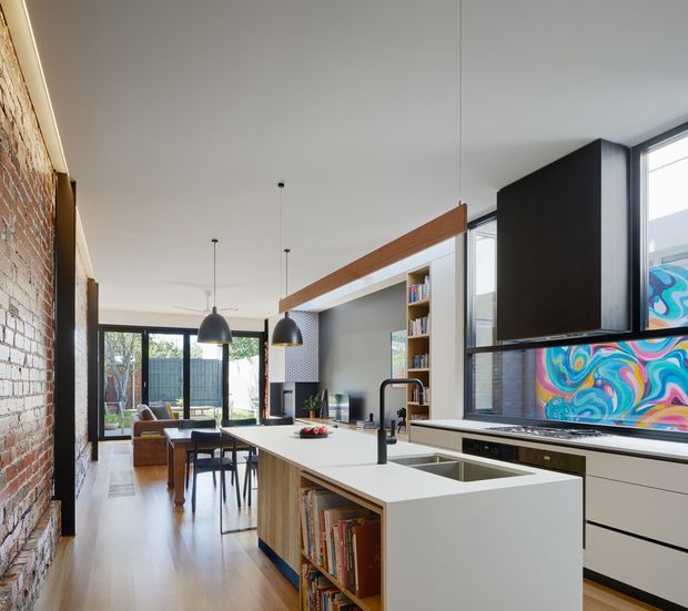 Open-plan contemporary kitchen and living area in a narrow, elongated interior. The kitchen features a large white island with integrated sink, pale timber cabinetry and open book shelving, dark lower cabinetry, a black-framed window wall, and a colourful abstract splashback or artwork behind the cooktop. Exposed red brick runs along the left wall, while timber flooring, pendant lights, and a visible dining and lounge area extend toward the rear. The space combines crisp modern joinery with warm timber and industrial brick detailing.