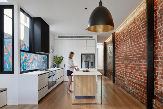 A narrow contemporary kitchen with a central island, white cabinetry, integrated appliances, and a black range hood. Floor-to-ceiling glazing and colourful patterned glass panels line one side, while an exposed red-brick wall creates a strong industrial feature. Timber flooring, dark joinery, and a large black-and-gold pendant add contrast.