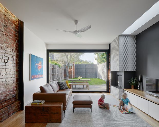 A bright contemporary living room with an exposed red-brick feature wall, brown leather sectional sofa, timber furniture, and a built-in black-and-white media wall with fireplace and television. Full-width glazing opens to a small lawn and fenced courtyard, while a large skylight and ceiling fan add light and ventilation. Children are playing on a pale rug in the foreground.