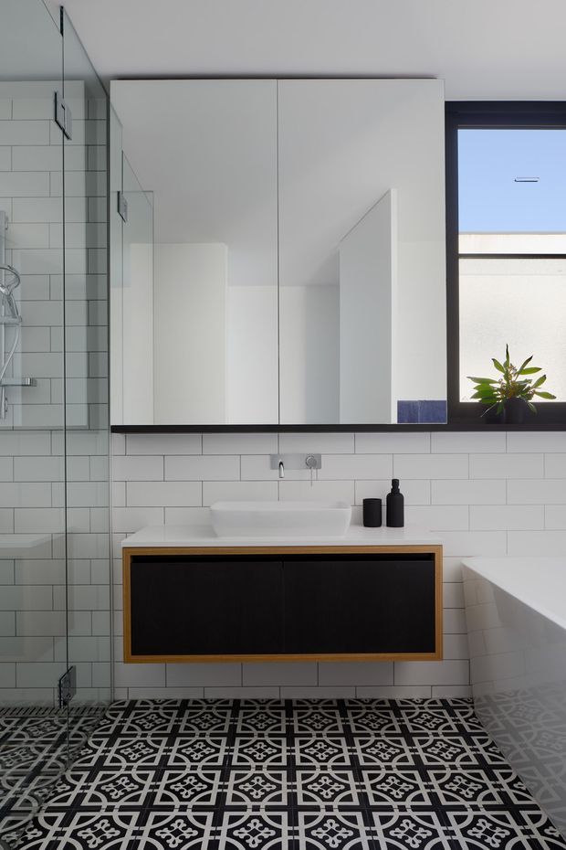 A crisp contemporary bathroom with a floating dark vanity framed in timber, white vessel basin, large mirrored wall cabinet, white subway-tile walls, and a bold black-and-white patterned floor. A frameless glass shower is visible on the left, while a white bathtub and black-framed window sit to the right. The palette is monochromatic with warm timber detailing and strong geometric contrast.