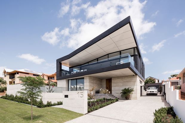 Exterior view of a large contemporary multi-level residence with a dramatic asymmetrical sloping roofline, expansive glazed balcony, dark metal framing, pale stone or concrete wall surfaces, and a broad driveway leading to a gated garage. The landscaped front garden and neighbouring homes reinforce a suburban setting.