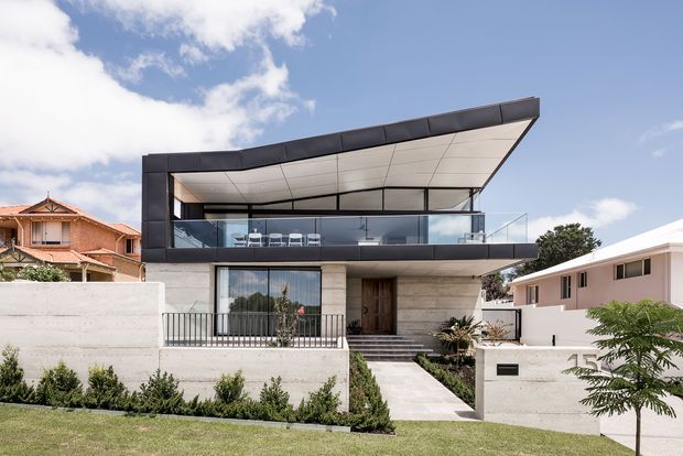 Exterior photograph of a contemporary multi-level residence with a dramatic cantilevered, angular roof canopy and dark metal-clad frame. The home has expansive glazed upper-level living space with outdoor seating, pale stone or concrete lower walls, a broad stepped entry, balcony balustrades, and landscaped front grounds. Neighbouring suburban houses are visible on either side beneath a partly cloudy sky.