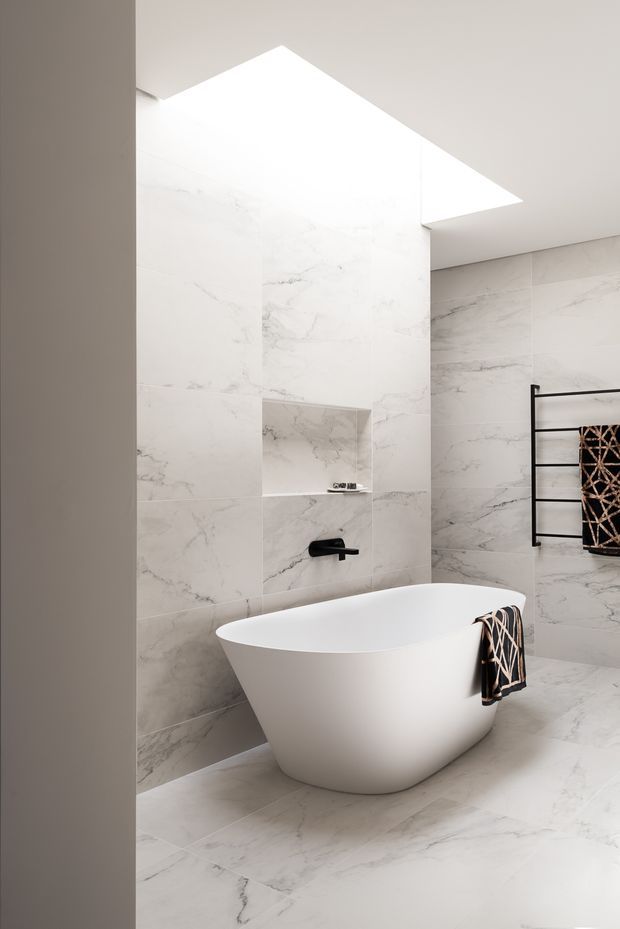 A bright, refined contemporary bathroom featuring full-height white marble-look wall and floor tiling with grey veining, a freestanding white oval bathtub, recessed wall niche, matte black wall-mounted tapware, and a black heated towel rail with patterned towels. A large angular skylight or ceiling opening provides abundant natural light.