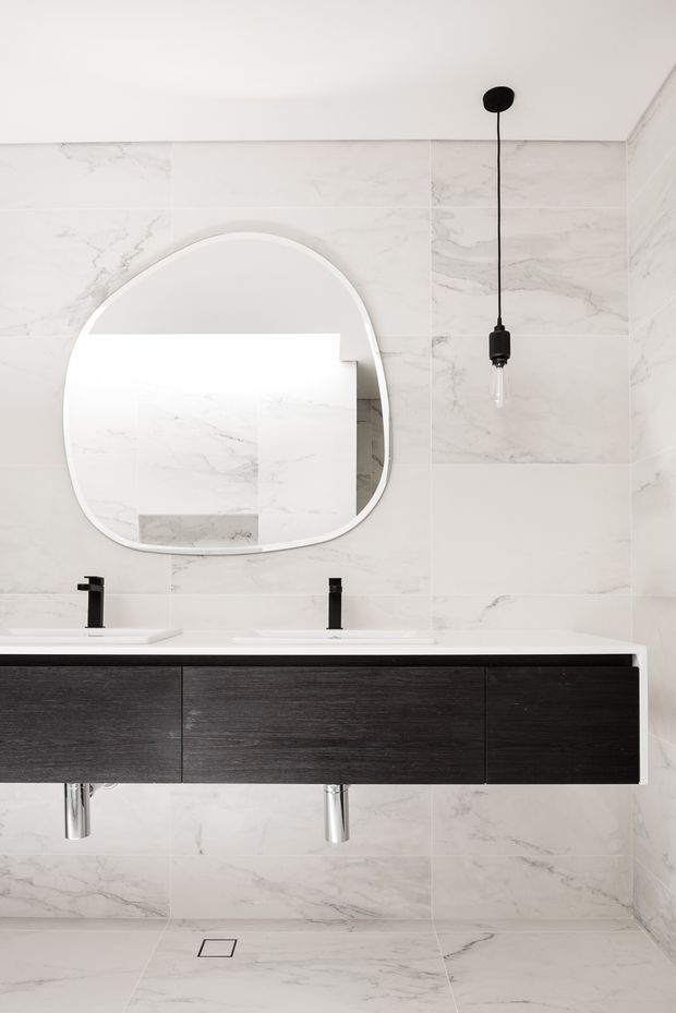 A contemporary bathroom featuring full-height white marble-look wall and floor tiles, a long floating dark timber vanity with two integrated white basins and black tapware, a large rounded mirror, and a minimalist black pendant light. The space is bright, clean, and restrained with strong monochromatic contrast.