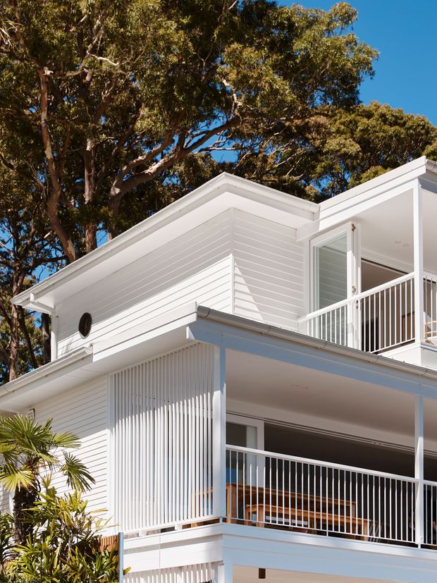 A bright white multi-level residential building exterior set among mature trees, featuring horizontal weatherboard cladding, crisp angular roof forms, open balconies with white metal balustrades, and a screened section of vertical battens. The architecture has a clean contemporary coastal character.