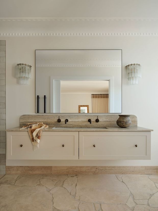 A refined bathroom vanity with a floating cream-painted double-drawer cabinet, weathered stone benchtop and integrated trough basin. Black wall-mounted tapware and handles contrast with the light joinery. A large rectangular mirror is framed by decorative wall moulding, with matching crystal wall sconces on either side. The room has pale limestone-style flooring and a warm, neutral palette with traditional detailing.