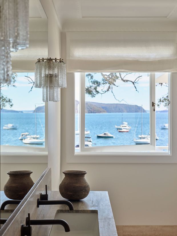 A bright, refined bathroom overlooking a blue waterfront through large white-framed windows. The room features a double vanity with a pale stone benchtop, undermount sinks, dark metal tapware, decorative pottery, a crystal pendant light, white panelled walls and ceiling, and a soft Roman blind. Boats and a wooded shoreline are visible outside.