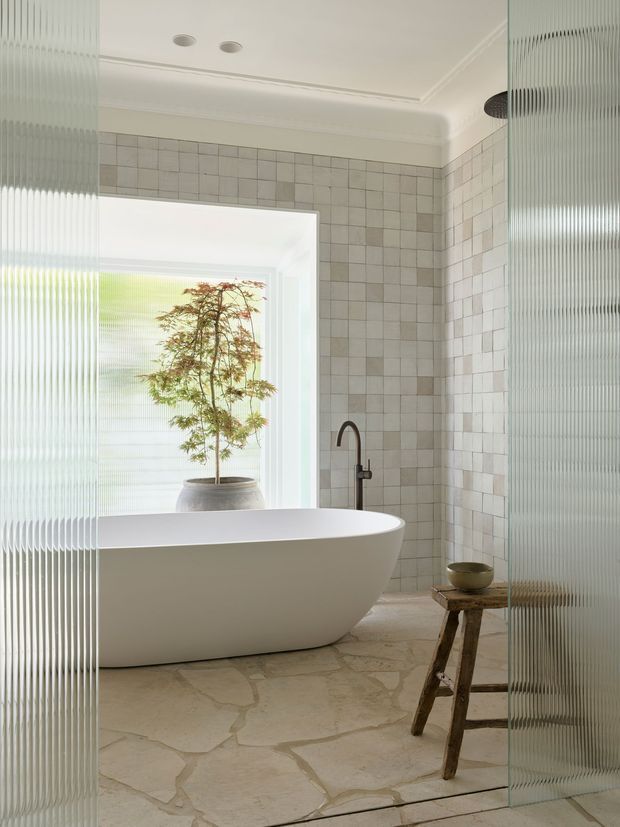A bright, refined bathroom featuring a freestanding white oval bathtub, pale square wall tiles, irregular natural stone flooring, and a dark metal wall-mounted bath filler. Ribbed translucent glass panels frame the view and provide privacy, while a large frosted-glass opening brings in soft daylight and overlooks a potted tree. The ceiling has crisp moulded detailing and recessed lights, creating a calm contemporary interior with traditional decorative touches.