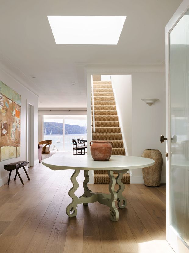 Bright, refined residential entry hall with a round sculptural console table in the foreground, pale timber flooring, white walls and trim, a carpeted staircase, and a large skylight. A dining and living area with expansive glazing and a water view is visible beyond the hallway. The space combines traditional architectural detailing with eclectic, artistic furnishings.