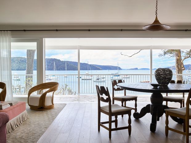 A bright, elegant dining and living area opening onto a waterfront view. The room features wide floor-to-ceiling glazing, pale timber flooring, a dark carved timber dining table with upholstered wooden chairs, curved rattan lounge chairs, a fringed rug, and a sculptural pendant light. A balcony or terrace with a metal balustrade overlooks a marina, hills, and calm water.