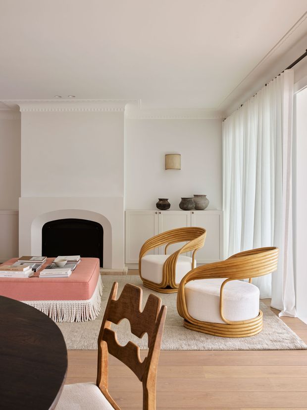 A bright, refined living room with white walls, ornate decorative cornice and fireplace detailing, full-height sheer curtains, pale timber flooring, and a neutral textured rug. Curved woven timber lounge chairs with white cushions surround a dark round dining table, while a pink upholstered ottoman with fringed trim sits near the fireplace. A white built-in sideboard displays ceramic vessels.
