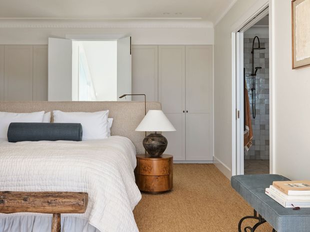 A refined, softly lit bedroom with a large upholstered bed, white textured bedding, built-in pale cabinetry, a rounded timber bedside table, and a sculptural table lamp. A bathroom with tiled walls and a shower is visible through the open doorway on the right. The room has carpeted flooring, white walls with decorative ceiling cornice, and a muted neutral palette with warm timber accents.