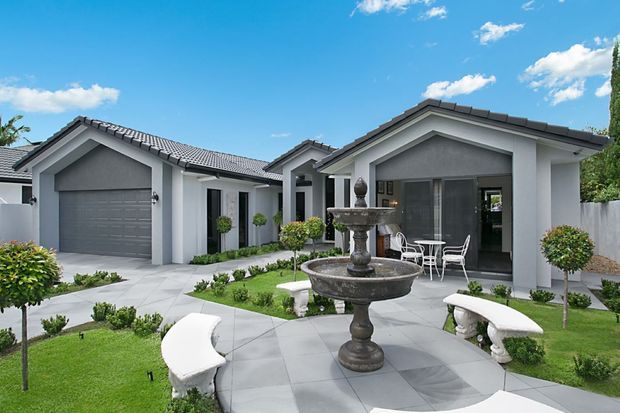Exterior view of a single-storey contemporary residence with white rendered walls, dark grey tiled gable roofs, an attached garage, and a landscaped courtyard. The front garden features geometric paving, clipped shrubs, curved white benches, and a central tiered stone fountain. A covered outdoor sitting area with glass doors is visible at the entry.