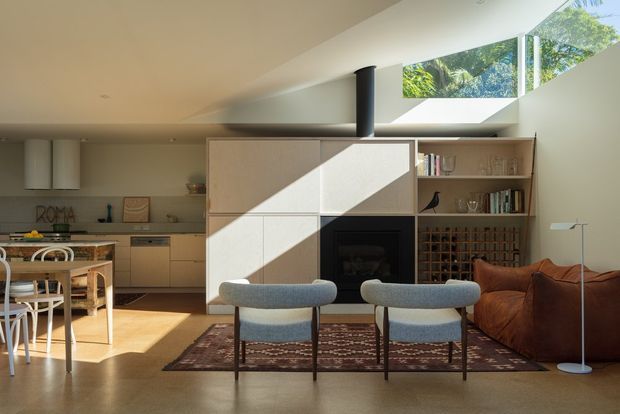 Open-plan contemporary living and kitchen space with a built-in central fireplace and shelving unit dividing the areas. The room features warm timber-toned flooring, pale built-in cabinetry, a dining table with chairs, a brown leather sofa, upholstered lounge chairs, and a patterned rug. Strong sunlight enters through a high clerestory-style glazed opening and skylight, creating a bright geometric light pattern across the fireplace wall.