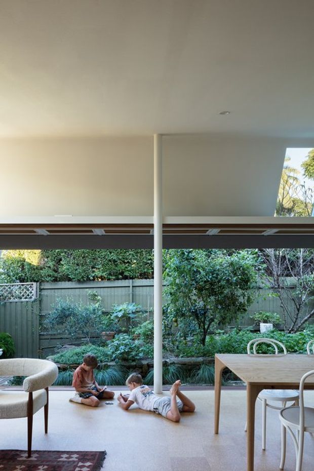A bright, open-plan interior with a covered outdoor connection and garden outlook. The room has a smooth white ceiling with recessed lighting, a central round support column, expansive glazing or open panels, and a pale floor. A light timber dining table with white chairs occupies the right side, while a curved upholstered lounge chair and patterned rug sit at left. Children are relaxing on the floor, and dense greenery is visible beyond the opening.