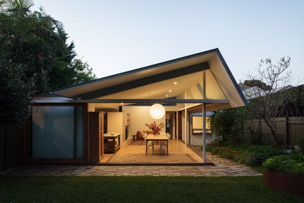 A compact modern pavilion-style home extension opening onto a paved garden terrace. The building has a dramatic asymmetrical pitched roof with broad eaves, extensive glazing, dark timber or metal framing, warm timber interior finishes, and a lit dining and living area visible through the open facade. Lush planting surrounds the structure, creating an indoor-outdoor connection.