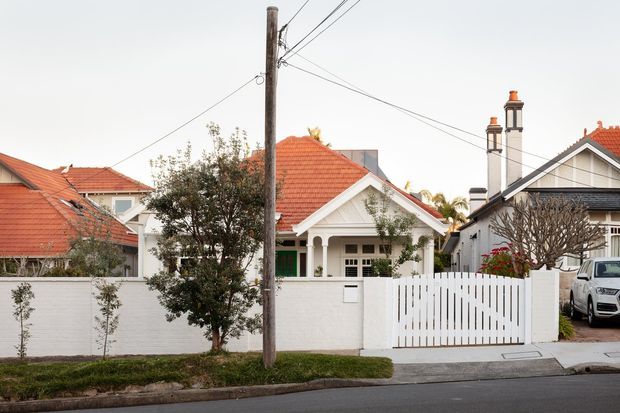 Street-level view of a suburban residential property featuring a single-storey white-rendered house with a prominent red terracotta tiled gabled roof, front porch, white masonry boundary wall, vertical timber-style gate, landscaped verge, and a visible driveway. Adjacent homes share similar pitched tiled roofs, with overhead utility wires and poles crossing the streetscape.