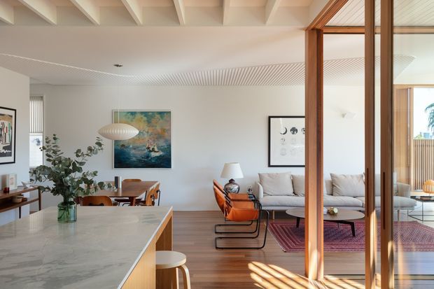 Open-plan residential interior connecting a kitchen, dining area and living room. A large pale stone kitchen island occupies the foreground, with timber cabinetry and stools. The dining area includes a timber table and chairs beneath a sculptural pendant light, while the living area has a light sofa, orange lounge chair, timber flooring and a patterned rug. Warm timber-framed glazing and doors divide the living space from a bright adjoining area. The ceiling combines exposed painted beams with a finely slatted section, creating a refined mid-century-inspired interior with abundant natural light.