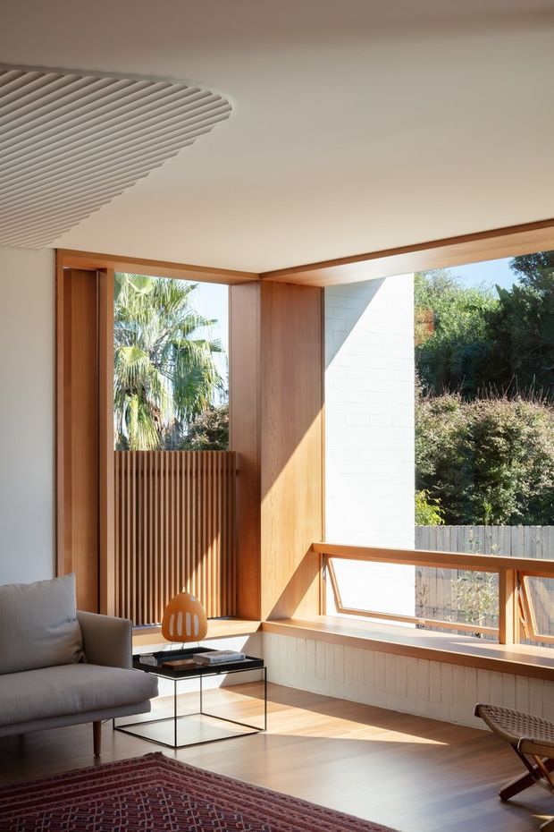 A sunlit mid-century-inspired living room with warm timber-framed corner glazing, a white masonry wall, timber flooring, and a sculptural ribbed ceiling detail. The room includes a neutral upholstered lounge chair, a small glass-and-black-metal side table, a patterned red rug, and a low timber-framed window seat or balustrade overlooking dense greenery. Strong natural light creates geometric shadows across the timber surfaces.