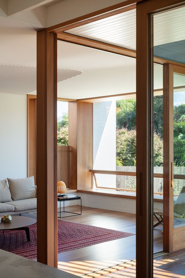 A bright, warmly detailed living room viewed through open timber-framed glazed doors. The space features extensive natural timber framing and joinery, pale walls, broad windows, a light timber floor, a large patterned red rug, a neutral sofa, and a small black coffee table. Sunlight filters through the glazing, with landscaped greenery visible outside. Linear slatted ceiling details and strong indoor–outdoor visual connection add architectural interest.