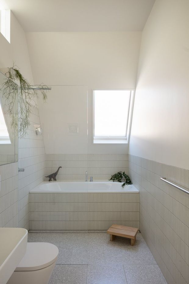 Bright, minimalist bathroom with a built-in white bathtub beneath a skylight, surrounded by fine vertically ribbed pale tiles. A partial toilet and vanity are visible in the foreground, with a frameless glass shower screen at left, a chrome grab rail, terrazzo-like flooring, and sparse plant and wooden stool accents.